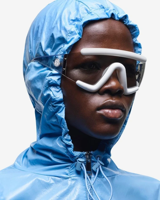 A model with a dark complexion wearing a light blue hooded jacket and distinctive white eyewear against a white backdrop.