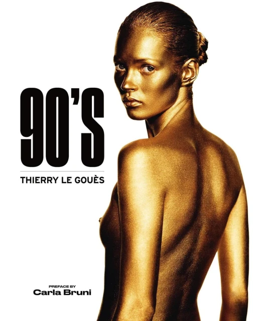 Gold-painted topless woman poses in front of white background with text. Fashion, beauty, and photography are the dominant attributes.