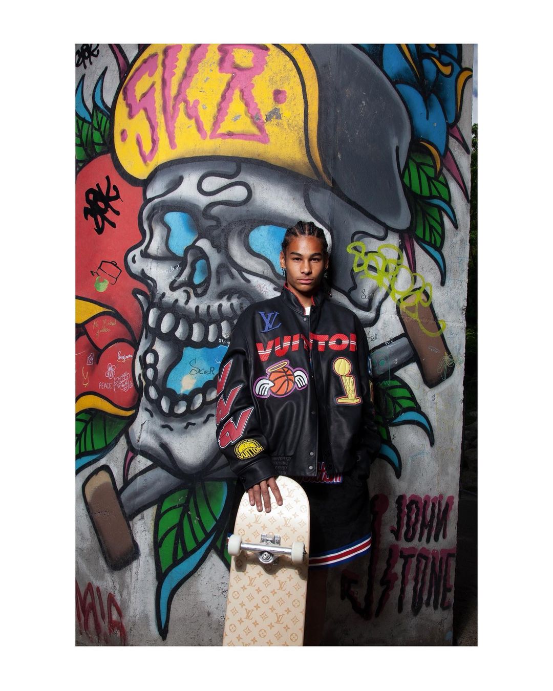 Teen poses with skateboard in front of graffiti art of skull, rose and crown, wearing Louis Vuitton logo jacket and black and white shorts.