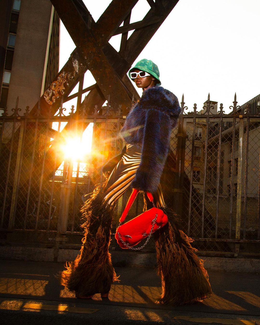 A fashion shot of a stylish adult model wearing flared pants, a furry jacket, and a green bucket hat in an urban outdoor setting.