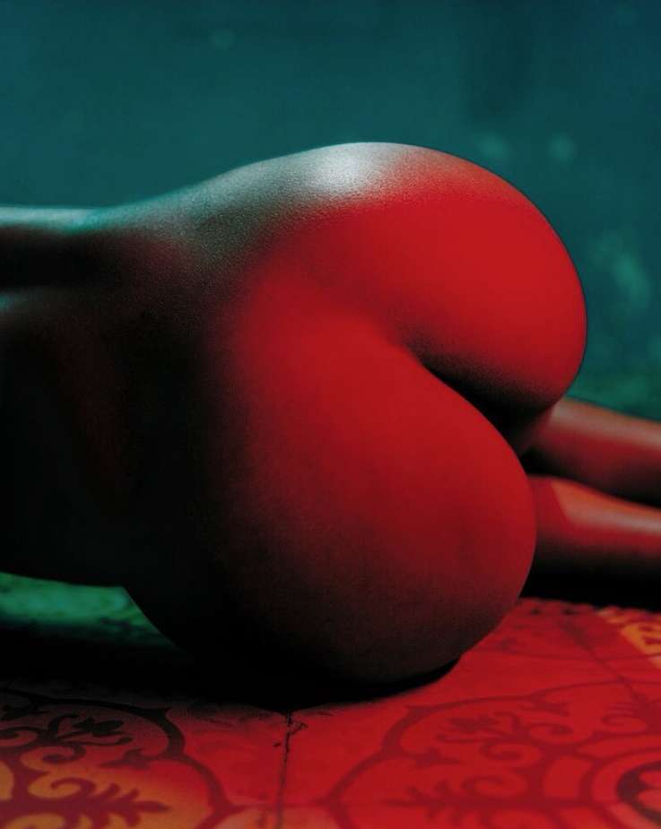 Close-up studio shot of an adult female with red lighting, showing her back and buttocks, lying on patterned fabric.