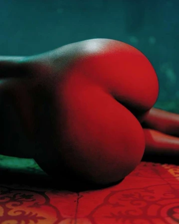 Close-up studio shot of an adult female with red lighting, showing her back and buttocks, lying on patterned fabric.