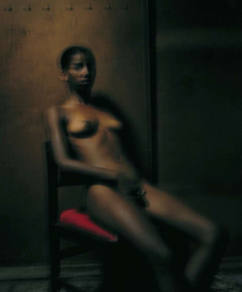 An adult woman sits in a chair with a red cushion, looking forward in a dimly lit interior with an aged wall behind her.