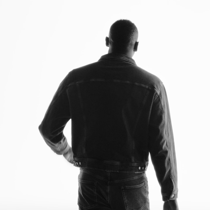 Monochrome portrait from behind of an adult man with short hair in a denim jacket and jeans in a brightly lit studio setting.