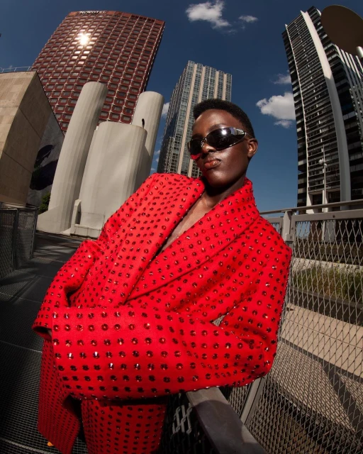 Adult female model wearing a red, embellished jacket and sunglasses stands outside against a backdrop of urban architecture and blue sky.