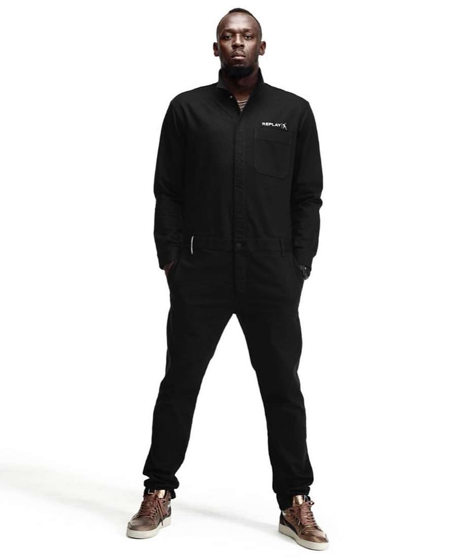 Full-length studio portrait of an adult male in a black jumpsuit and brown high-top shoes, striking a confident pose against a white backdrop.