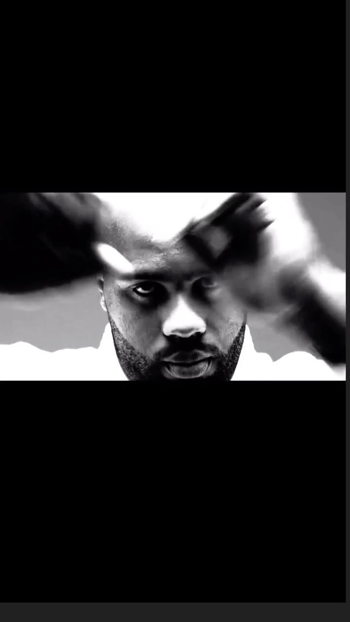 Close-up monochrome portrait of a bald Black man with a beard looking directly forward with motion-blurred hands.