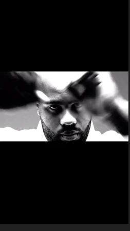 Close-up monochrome portrait of a bald Black man with a beard looking directly forward with motion-blurred hands.