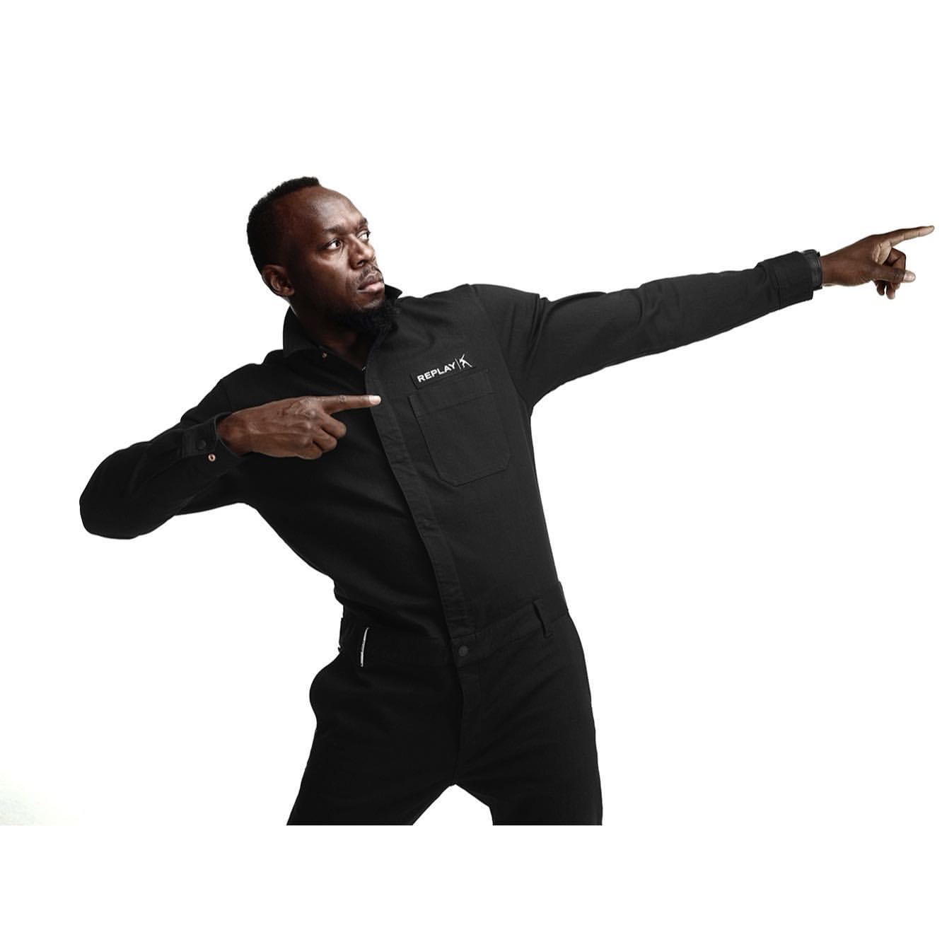 Adult male in black jumpsuit with arms outstretched pointing in studio. Solid white background. Brand logo visible on shirt.