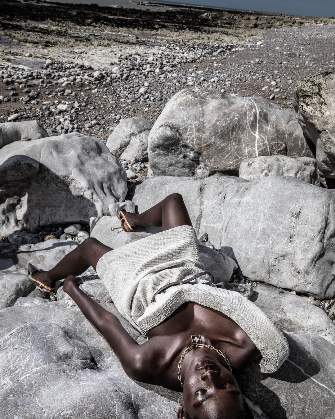 Adult female model poses on rocky beach in a light-colored dress and gold jewelry; natural light, dark skin, rocks, sand, and ocean.
