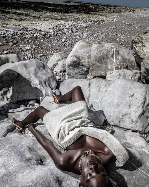 Adult female model poses on rocky beach in a light-colored dress and gold jewelry; natural light, dark skin, rocks, sand, and ocean.