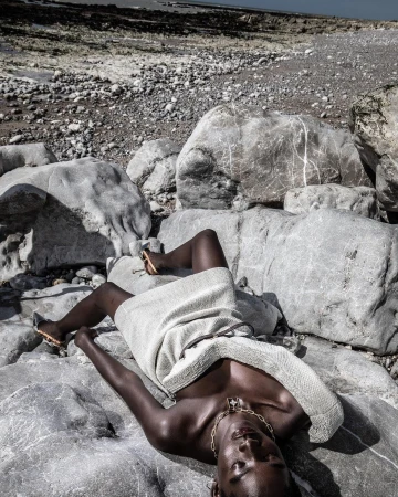 Adult female model poses on rocky beach in a light-colored dress and gold jewelry; natural light, dark skin, rocks, sand, and ocean.