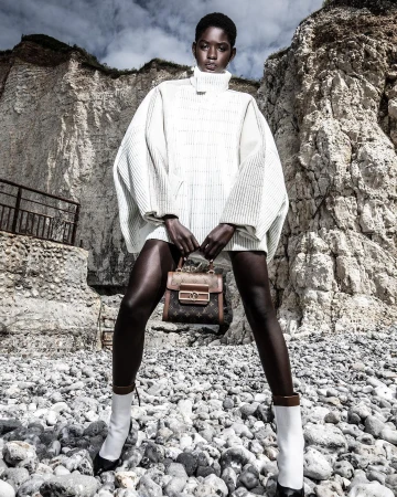 Fashion shot of an African woman posing with a purse on a rocky beach wearing a white top, with cliffs in the background.