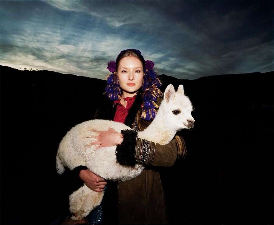 A young woman in traditional clothing holds a white alpaca against a dark, mountainous backdrop under an overcast sky.
