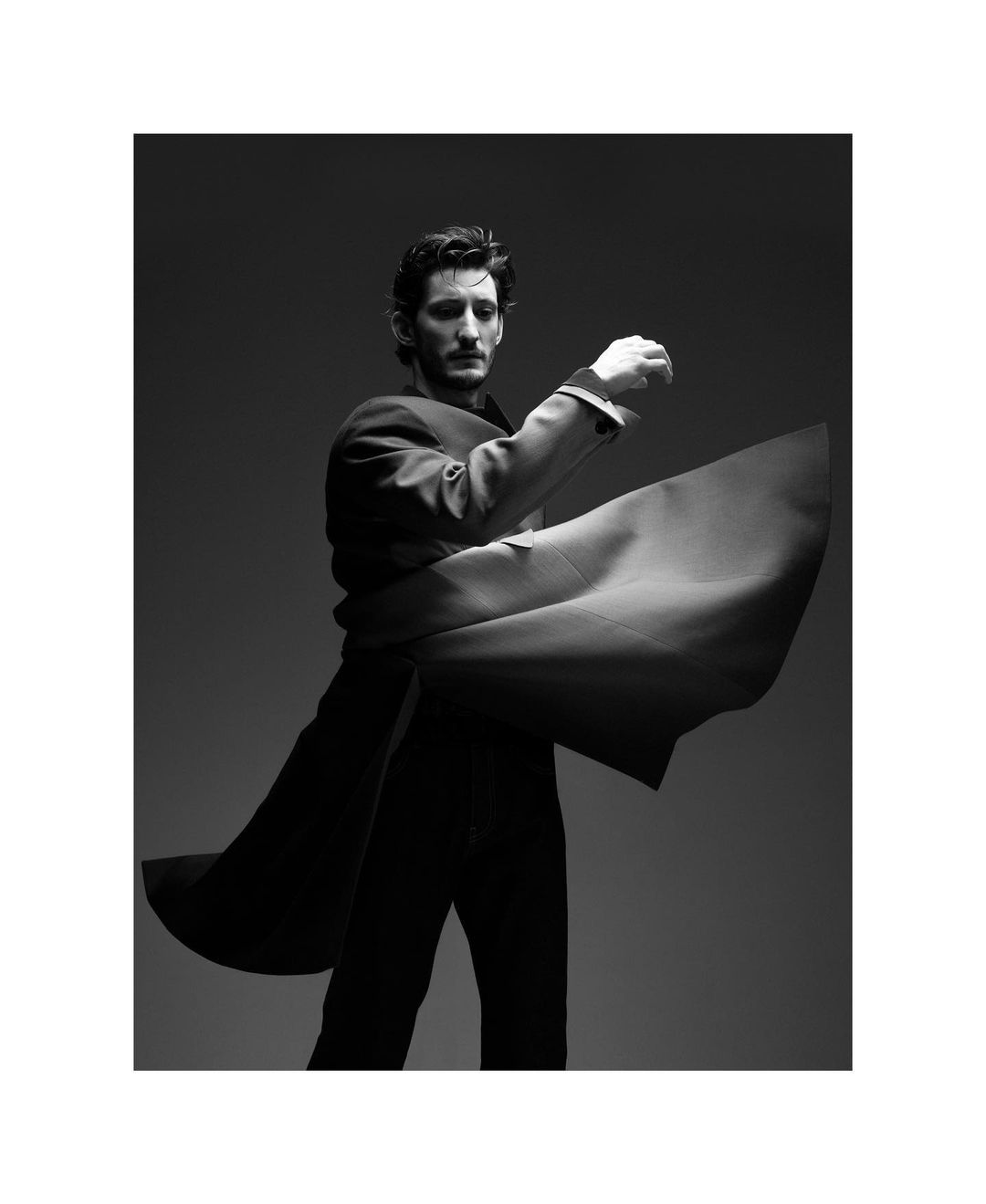Black and white fashion shot of an adult man in a suit coat with a flowing garment wrapped around him, posed against a grey backdrop.