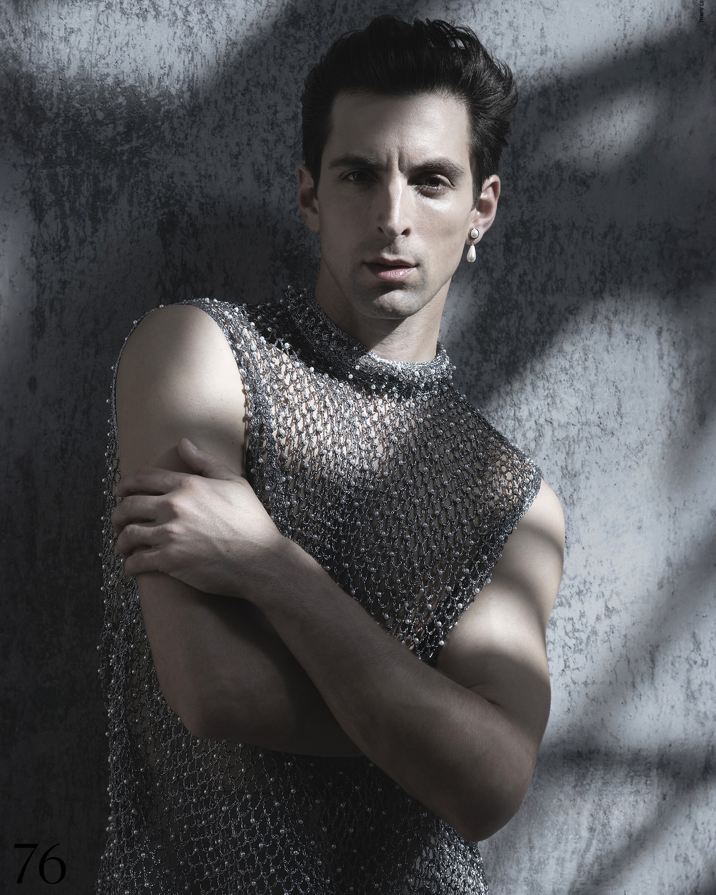 Man in beaded mesh top poses in front of textured grey wall with dappled light. Portrait orientation, studio lighting, neutral facial expression.