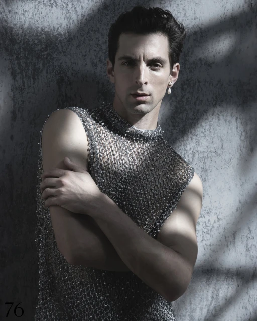 Man in beaded mesh top poses in front of textured grey wall with dappled light. Portrait orientation, studio lighting, neutral facial expression.
