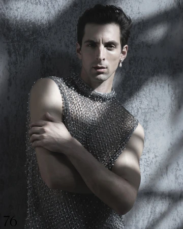 Man in beaded mesh top poses in front of textured grey wall with dappled light. Portrait orientation, studio lighting, neutral facial expression.