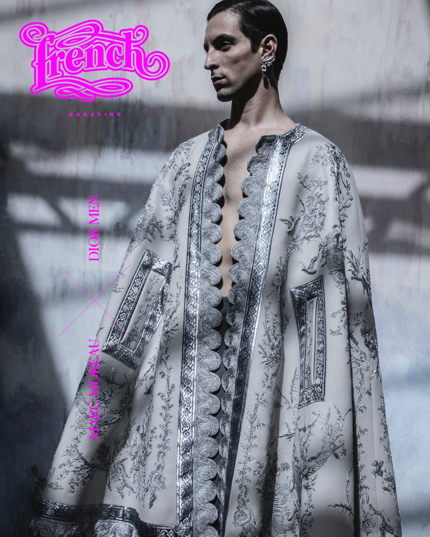 Fashion magazine cover featuring a male model wearing a cape with an ornamental pattern against a neutral background with diffused light.