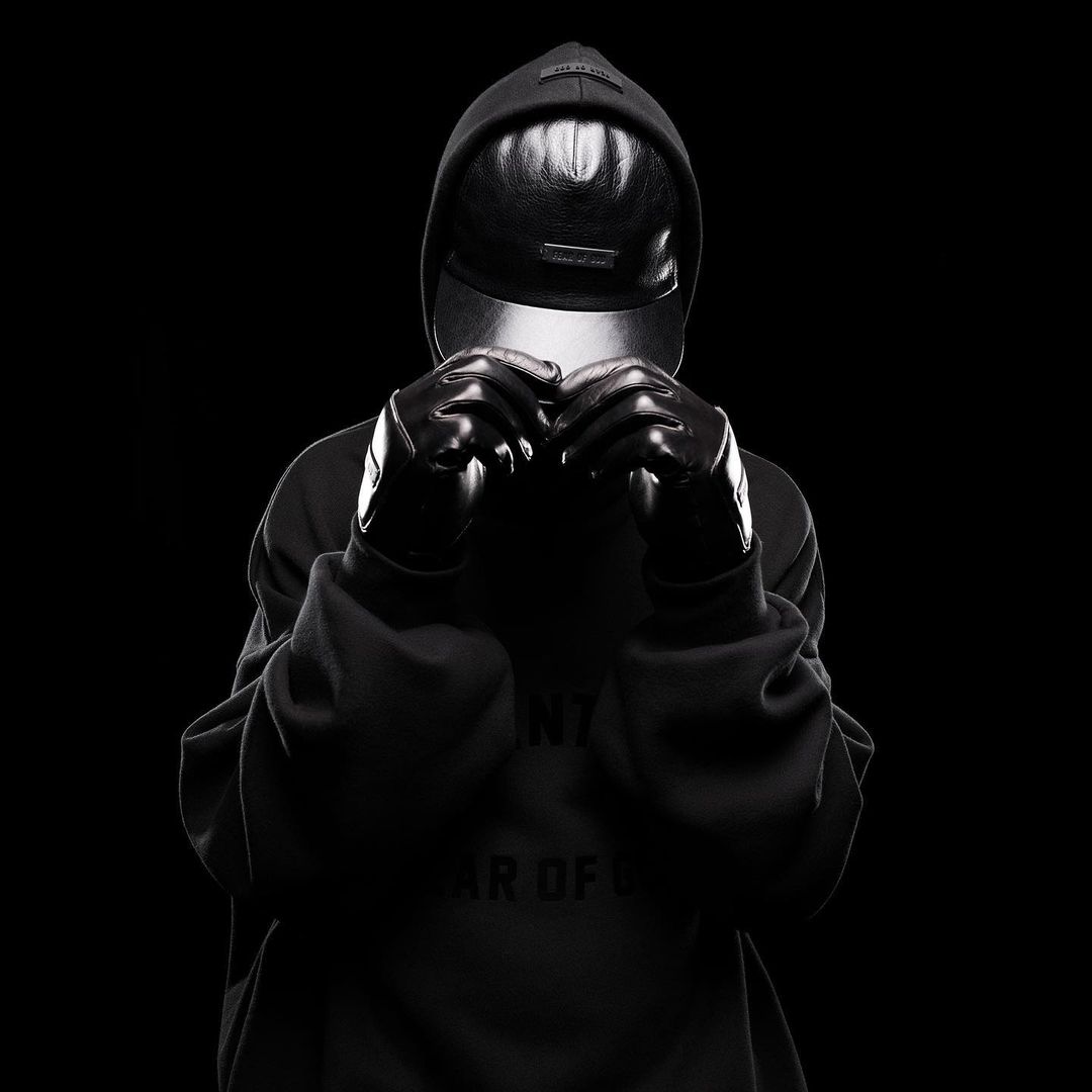 Person in hooded sweatshirt and gloves in a studio environment. The person is obscured by a visor and dark gloves against a black background.