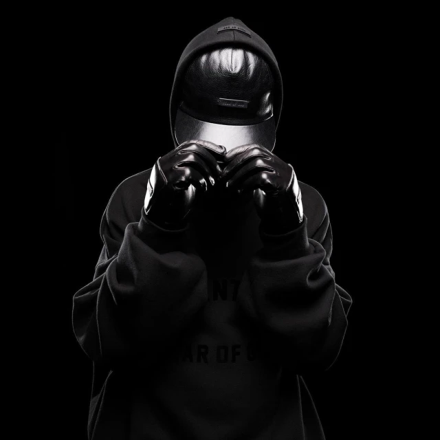 Person in hooded sweatshirt and gloves in a studio environment. The person is obscured by a visor and dark gloves against a black background.