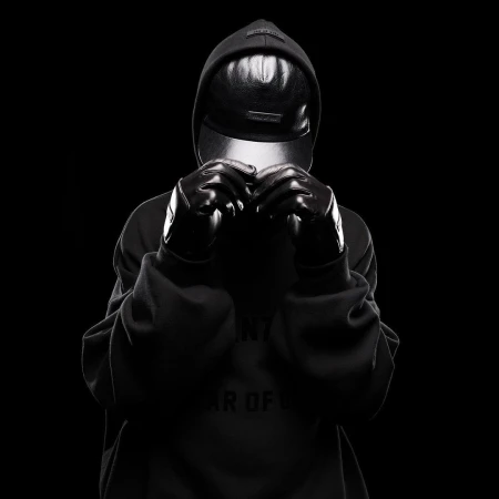 Person in hooded sweatshirt and gloves in a studio environment. The person is obscured by a visor and dark gloves against a black background.