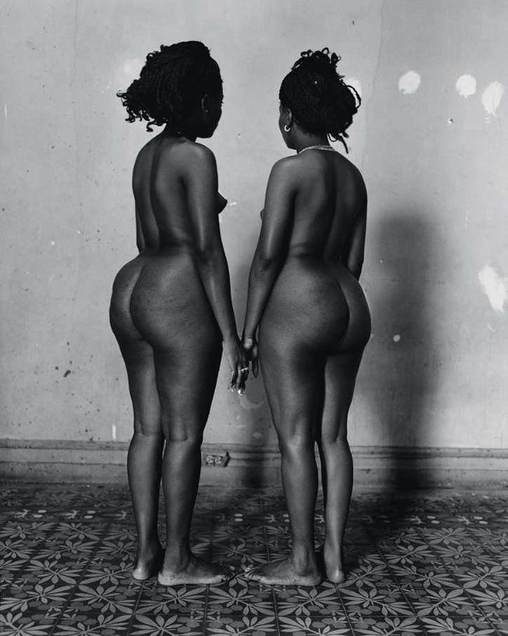 Black and white portrait of two nude Black adult females standing closely together in an indoor environment with patterned flooring.