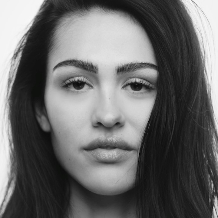 Striking black and white portrait of a young adult woman with long dark hair and neutral expression against a clean, bright background.