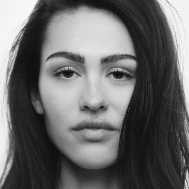 Striking black and white portrait of a young adult woman with long dark hair and neutral expression against a clean, bright background.
