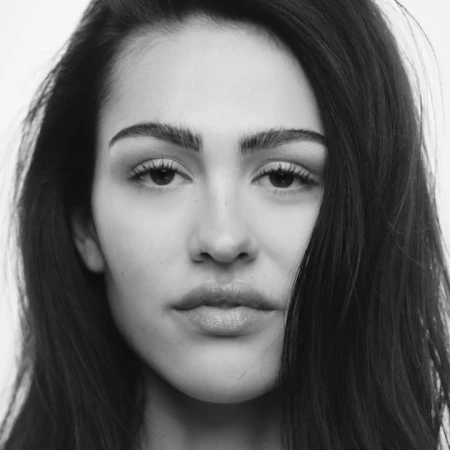 Striking black and white portrait of a young adult woman with long dark hair and neutral expression against a clean, bright background.