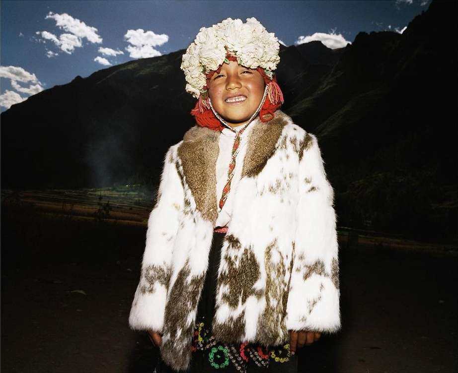 Smiling child in traditional clothing stands in a mountain landscape, wearing a flower crown and rabbit fur coat.