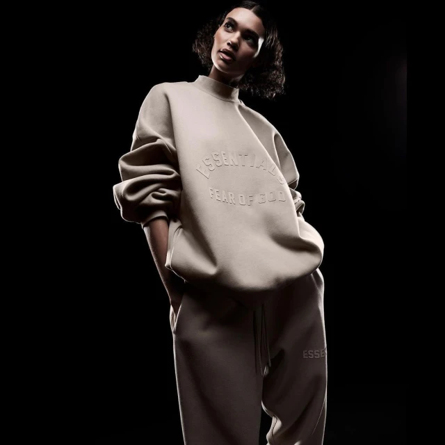 Woman poses in a studio wearing a beige 'Essentials Fear of God' sweatshirt and sweatpants against a dark background, low angle.