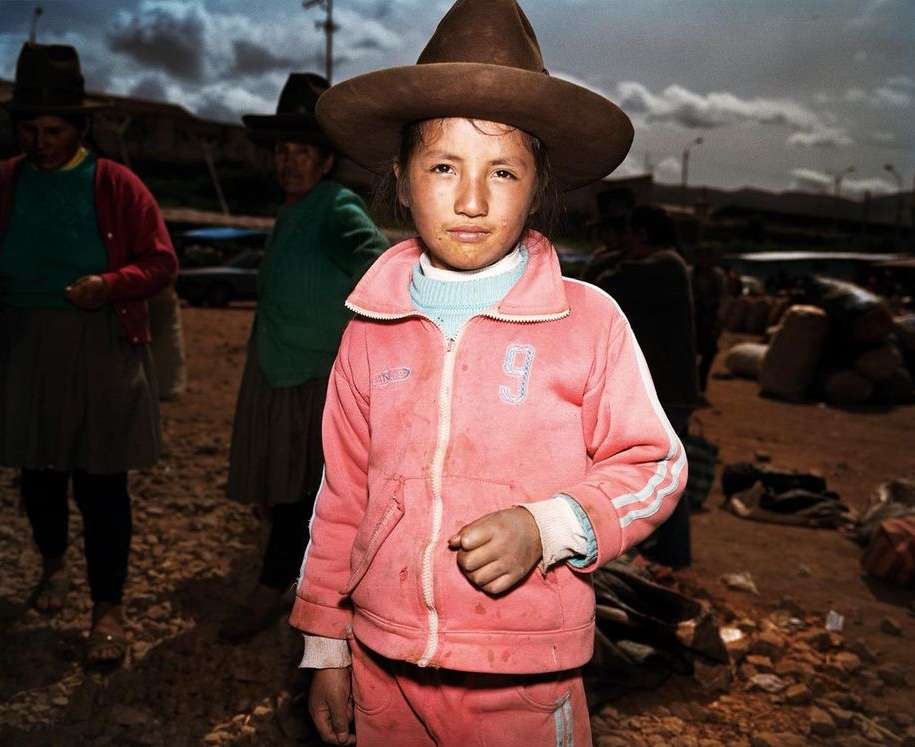 Young girl in pink jacket and hat stands in a crowded outdoor market area with other people, ground, and cloudy sky visible.