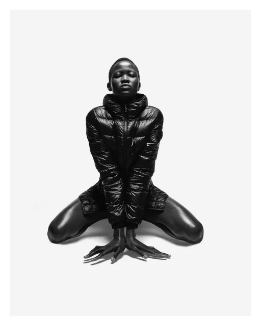 Monochromatic studio shot of an African female model kneeling and posing in a shiny black puffer jacket and shorts on a white background.