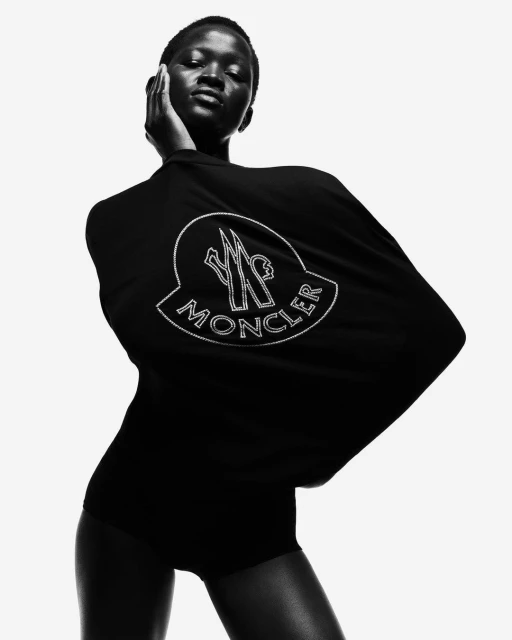 Close-up studio shot of a Black female model wearing a black bodysuit and a Moncler logo shirt, posing against a bright white backdrop.