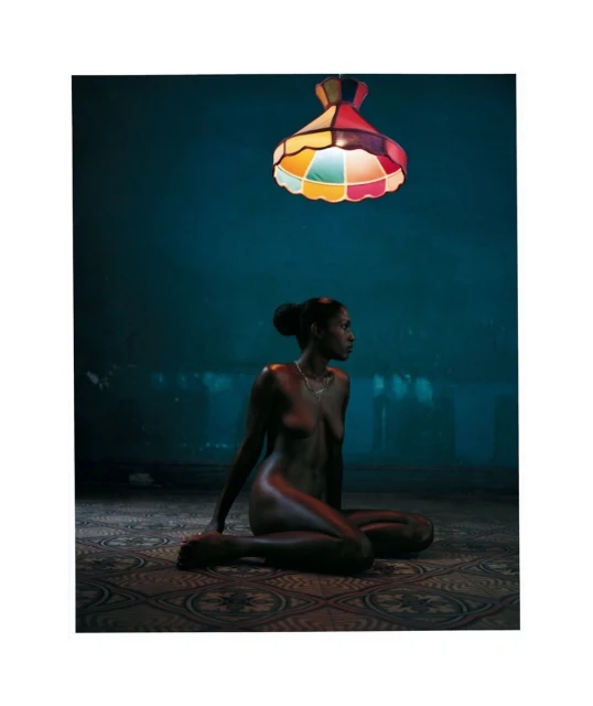 Bare-chested woman with dark skin in a seated pose beneath a colorful lamp, with a dark blue backdrop and patterned floor.
