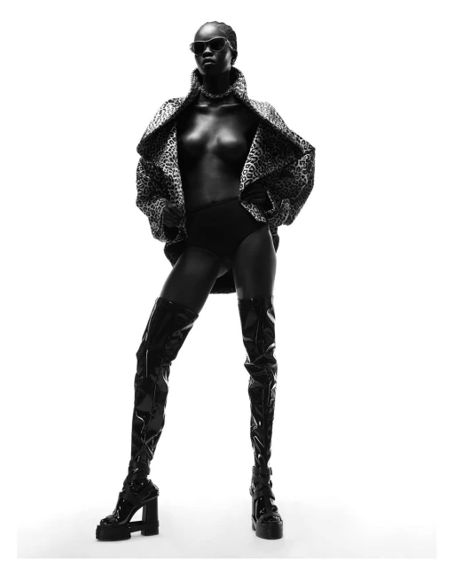 Monochrome studio shot: Black woman in sunglasses and thigh-high boots poses in an open leopard-print jacket against a white backdrop.