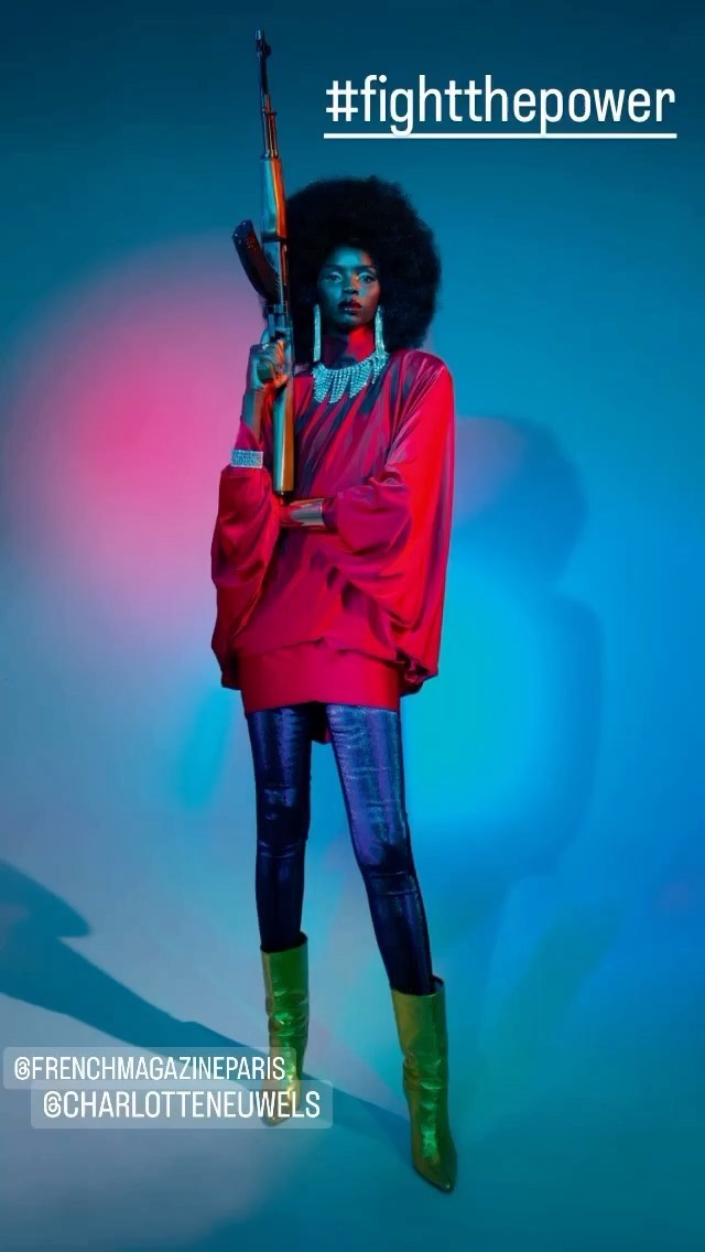 A black woman in red holds a rifle while posing in a studio with blue and pink lighting. She wears green boots and has an afro.