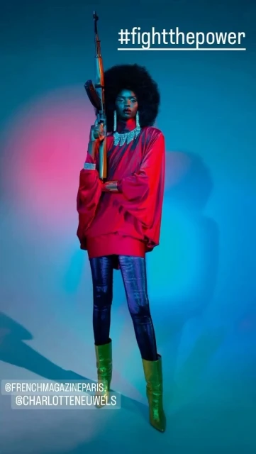 A black woman in red holds a rifle while posing in a studio with blue and pink lighting. She wears green boots and has an afro.