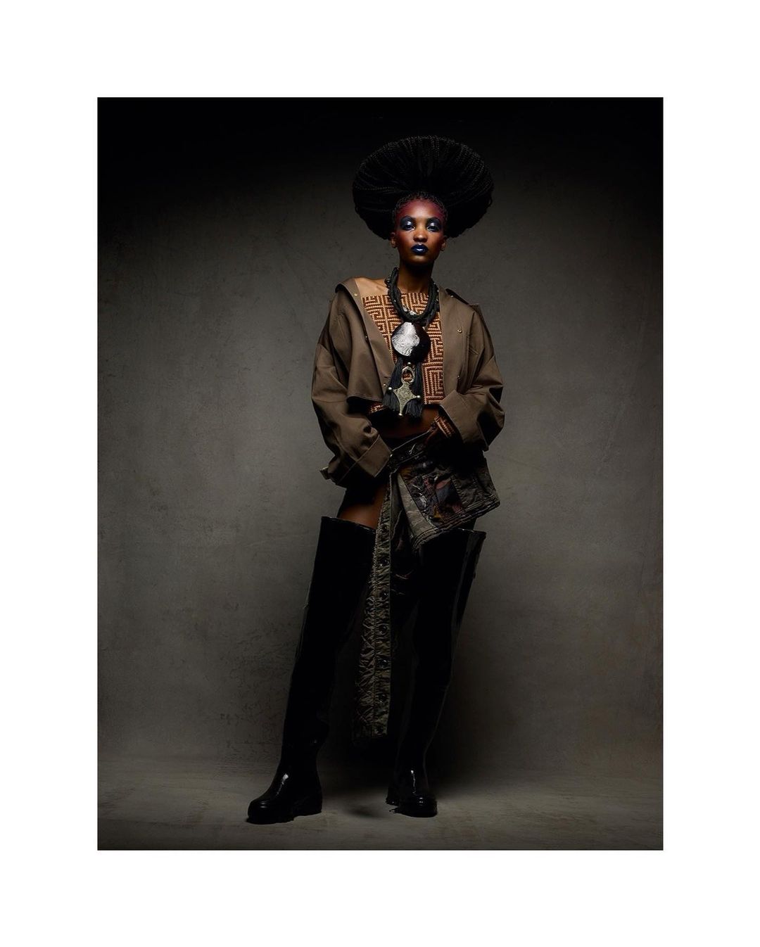 Adult female fashion model poses in a studio with makeup, a hat, and boots, wearing brown jacket and patterned material around the waist.