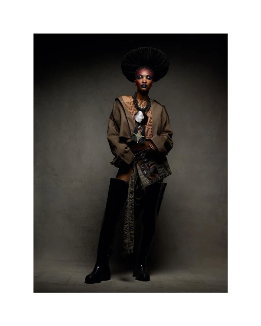 Adult female fashion model poses in a studio with makeup, a hat, and boots, wearing brown jacket and patterned material around the waist.