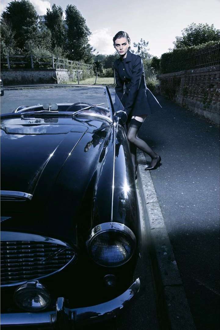An adult female in a dark coat poses next to a vintage black convertible car in an outdoor urban setting, wearing stockings and dark shoes.