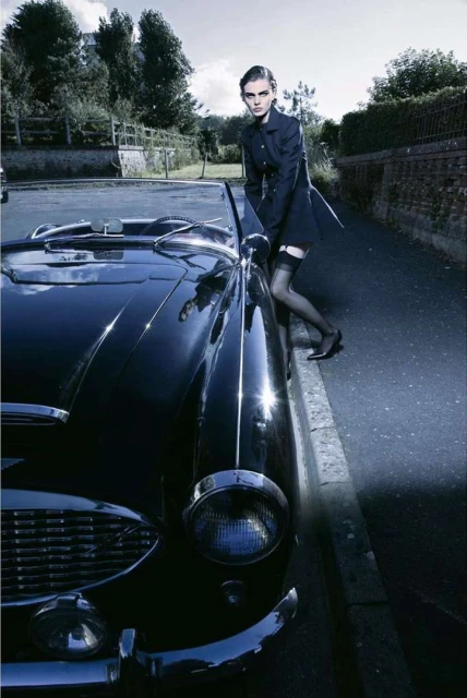 An adult female in a dark coat poses next to a vintage black convertible car in an outdoor urban setting, wearing stockings and dark shoes.