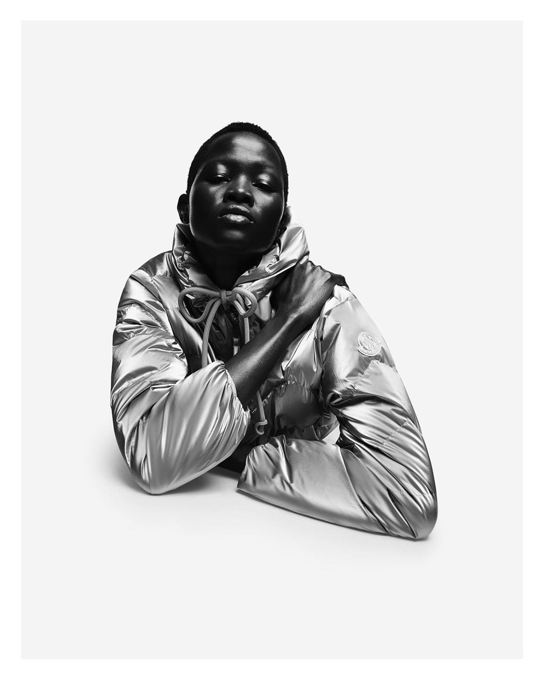 Close-up black and white portrait of a woman wearing a shiny, silver puffer jacket with a hood, set against a clean, bright white backdrop.