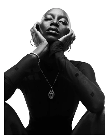 Portrait of a bald Black woman wearing a pendant necklace and sheer patterned top against a white background
