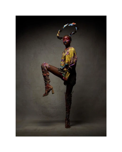 Stylish woman in patterned top and boots strikes a pose in a studio, with her leg raised and patterned scarf-like hair ornament.
