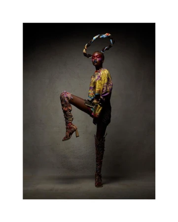 Stylish woman in patterned top and boots strikes a pose in a studio, with her leg raised and patterned scarf-like hair ornament.