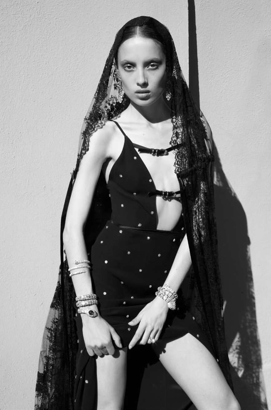 Black and white fashion portrait of a woman in a dress and lace veil, jewelry, standing against a textured wall in natural light.