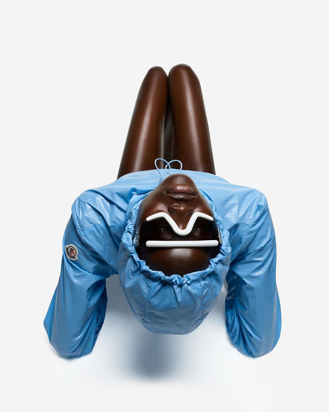 Dark-skinned fashion model in blue hooded jacket, lying upside down against white background. White stylized sunglasses frame eyes.