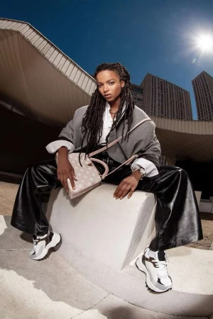 Black woman with long braided hair wearing wide black trousers and a grey jacket holding a patterned handbag outdoors against modern architecture.